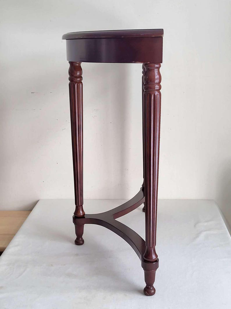 Op de afbeelding: Een donker bordeauxrode, halfronde bijzettafel met een driepoot. De tafel heeft een glad, gepolijst oppervlak en decoratieve, gefreesde poten. De tafel is ongeveer 76 cm hoog.