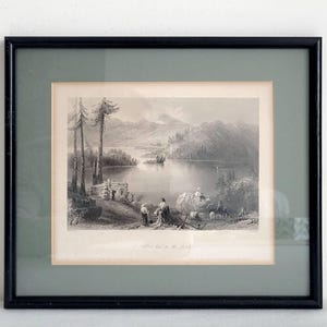 May include: A framed black and white landscape print showing a tranquil lake scene with figures, trees, and a small cabin. The print is set within a black frame and a light green mount.
