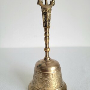 May include: A vintage brass bell with an ornate handle and detailed carvings. The bell has a flared base and a long handle with a decorative top. The brass has a warm, aged patina, suggesting its age and use. The bell is likely used for ceremonial or decorative purposes.