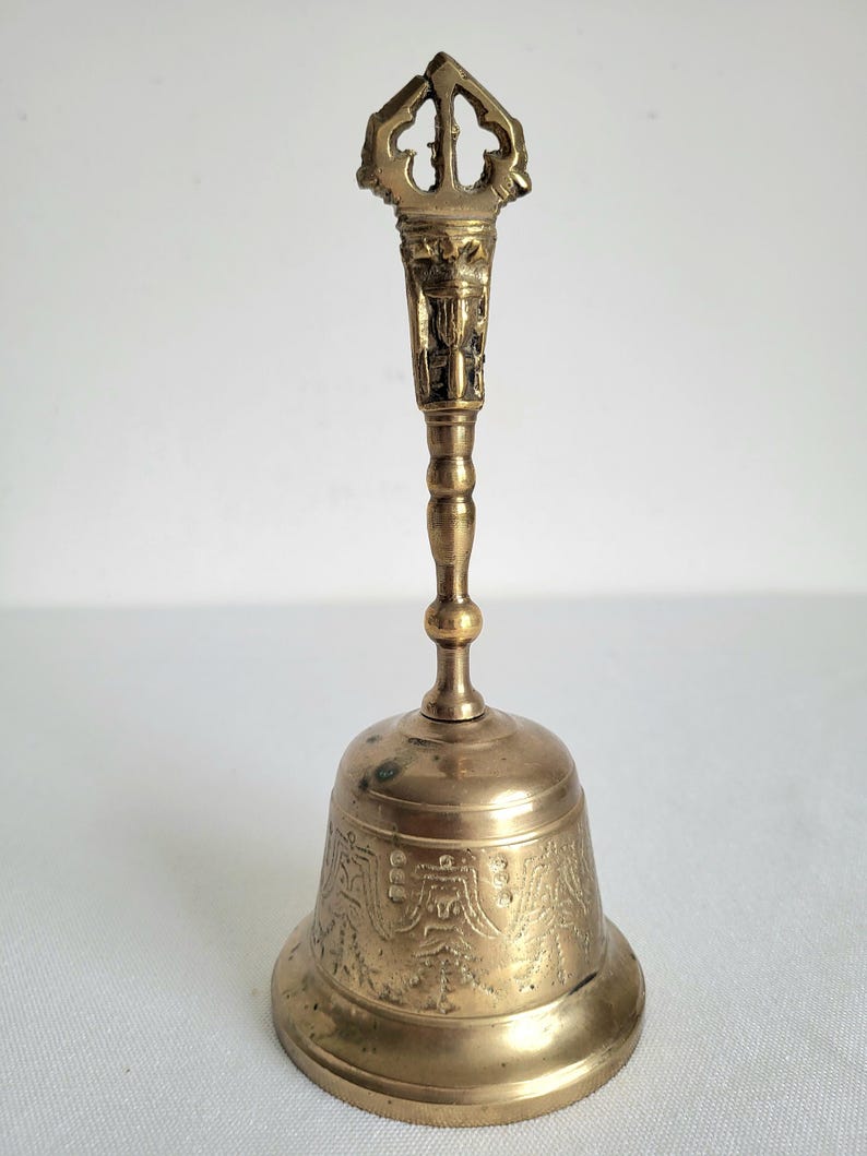 May include: A vintage brass bell with an ornate handle and detailed carvings. The bell has a flared base and a long handle with a decorative top. The brass has a warm, aged patina, suggesting its age and use. The bell is likely used for ceremonial or decorative purposes.