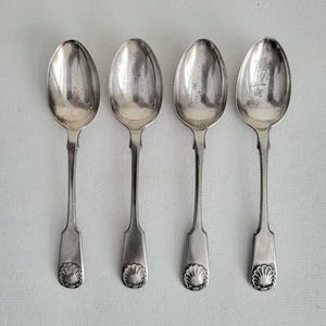 May include: Four antique silver spoons on a white background. Each spoon features an oval bowl, a long handle, and a shell design at the base. The spoons are reflective, showing signs of age and use. The spoons are made of silver.