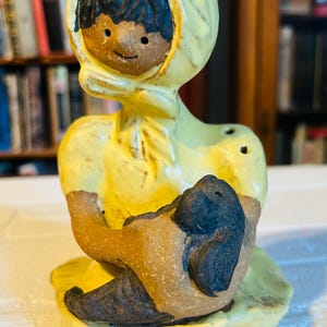 May include: A ceramic figurine of a girl wearing a yellow hooded dress and holding a small brown animal. The girl has dark hair and a smiling face.