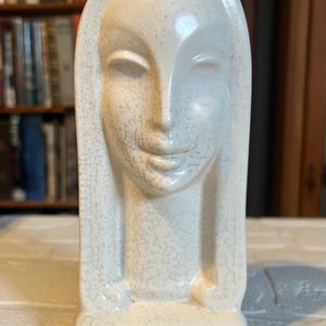 May include: A white ceramic sculpture of a woman's head with a stylized face and long hair. The sculpture has a crackled glaze finish.