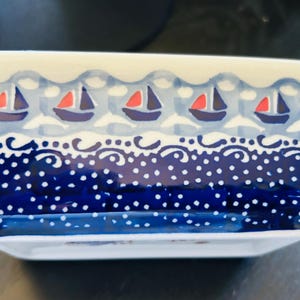 May include: A rectangular ceramic dish featuring a hand-painted pattern of small sailboats with red and blue sails on a light blue background, above a dark blue band decorated with white polka dots and swirling wave-like motifs along the top edge.