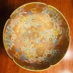 May include: A decorative bowl with a gold rim and a floral pattern in shades of gold, orange, blue, and white. The bowl's interior is covered in a detailed, repeating design of stylized flowers.