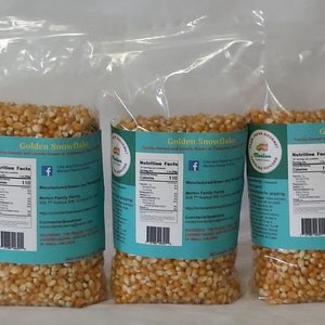 May include: Three clear plastic bags filled with golden popcorn kernels. Each bag has a teal label with the words "Golden Snowflake" and nutrition facts. The bags are sealed and ready for popping.
