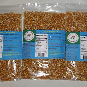 May include: Three clear plastic bags filled with golden popcorn kernels. Each bag has a blue label with the text "Golden Snowflake" and nutrition facts. The bags are labeled as 1 lb (16 oz).