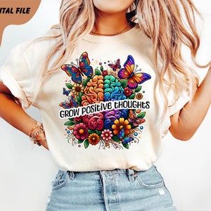 May include: A white t-shirt with a colorful brain design surrounded by flowers and butterflies. The text "GROW POSITIVE THOUGHTS" is printed on the shirt.