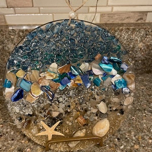 Könnte beinhalten: Ein rundes Dekorationsobjekt mit einer Strandszene. Der obere Bereich ist mit blauen und türkisfarbenen Glasstücken gefüllt, die in Muscheln und Sand übergehen. Ein Seestern und Muscheln sind sichtbar. Ein goldfarbener Ständer stützt das Objekt.