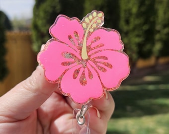 Puede incluir: Un portatarjetas con forma de flor de hibisco rosa brillante con un acabado de purpurina y un estambre amarillo central. La pieza floral de acrílico está unida a un mecanismo de cordón retráctil de plástico transparente con un clip metálico.
