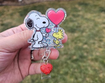 May include: A Snoopy and Woodstock themed badge reel. The badge reel features Snoopy holding a heart and Woodstock holding a pink heart-shaped balloon. A red heart charm is attached to the reel. The design is outlined with glitter.