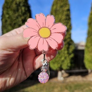 May include: A pink daisy-shaped badge reel with a yellow center and glitter accents. The badge reel has a clear plastic clip and a decorative pink beaded charm. The background features green trees and a blue sky.