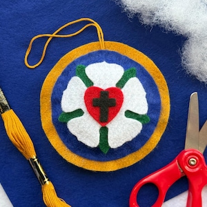 May include: A felt ornament with a white rose, green leaves, and a red heart with a black cross in the center. The ornament is surrounded by a blue circle with a gold border.
