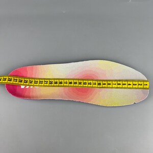 May include: A pink, yellow, and white insole is measured with a yellow measuring tape. The insole has a gradient design. The measuring tape shows measurements in inches, starting from 1 inch to 29 inches.