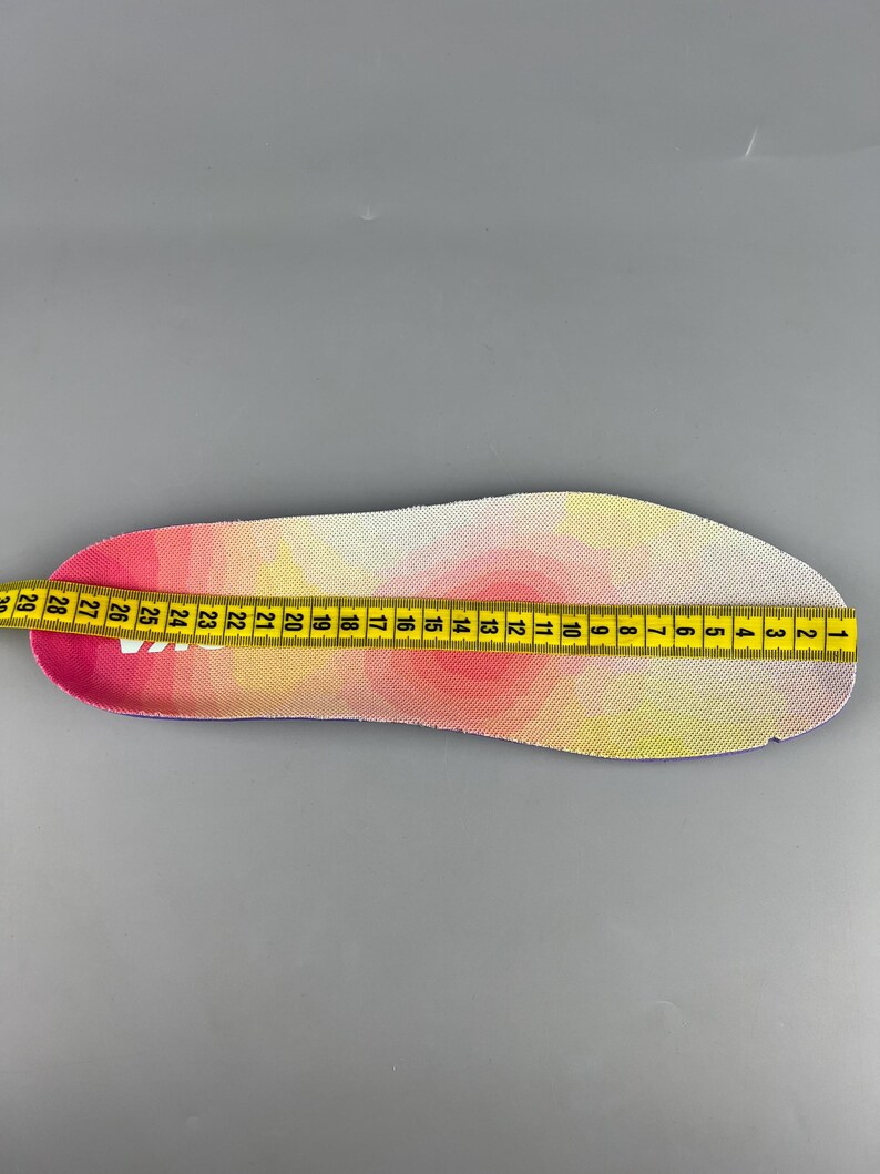 May include: A pink, yellow, and white insole is measured with a yellow measuring tape. The insole has a gradient design. The measuring tape shows measurements in inches, starting from 1 inch to 29 inches.
