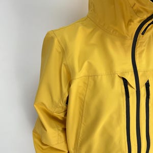 May include: A yellow hooded jacket with black accents. The jacket features a full-length zipper, multiple pockets, and a black panel at the bottom. The jacket is made of a smooth, water-resistant material.