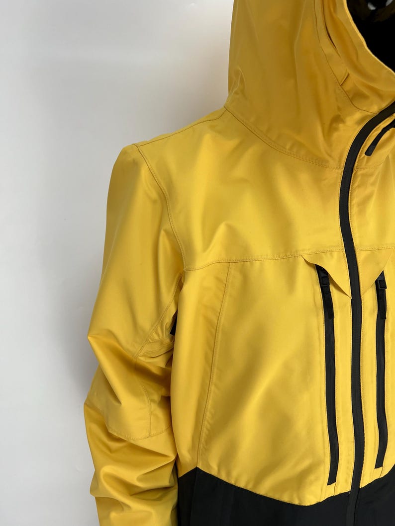 May include: A yellow hooded jacket with black accents. The jacket features a full-length zipper, multiple pockets, and a black panel at the bottom. The jacket is made of a smooth, water-resistant material.