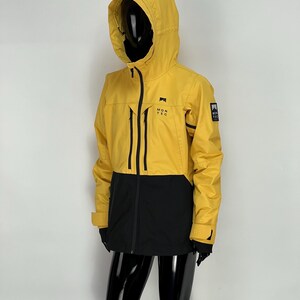 Puede incluir: Chaqueta con capucha amarilla y negra con el texto "MONTEC" en el pecho. La chaqueta tiene cremallera frontal, detalles negros y un pequeño parche con el logotipo en la manga. La chaqueta se muestra en un maniquí.