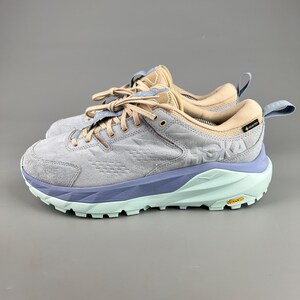 May include: Pair of gray, blue, and tan athletic shoes. The shoes have a light blue sole, a gray upper, and tan accents. The brand name "HOKA" is visible on the side. The shoes have laces and a pull tab on the heel.