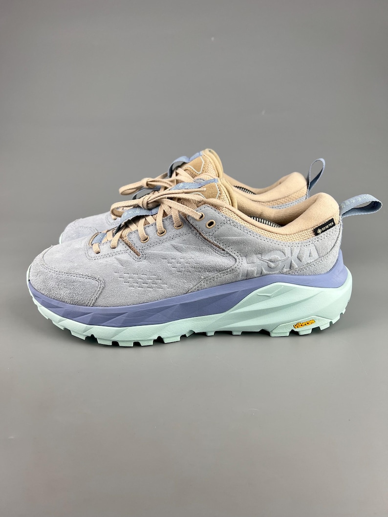 May include: Pair of gray, blue, and tan athletic shoes. The shoes have a light blue sole, a gray upper, and tan accents. The brand name "HOKA" is visible on the side. The shoes have laces and a pull tab on the heel.