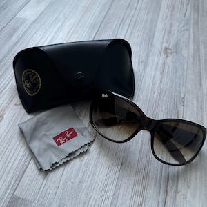 May include: Sunglasses with a dark brown frame and gradient lenses, alongside a black case and a grey cleaning cloth. The case features a circular logo. The cleaning cloth has a red rectangular logo.