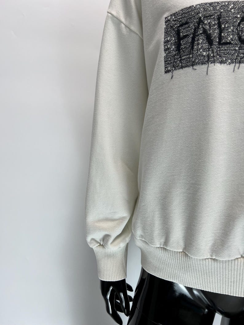 May include: Cream-colored sweatshirt with the word "FALC" in a textured, black font across the chest. The sweatshirt has long sleeves with ribbed cuffs and a ribbed hem. The fabric appears to be a soft, comfortable material.