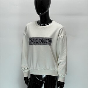 May include: A cream-colored sweatshirt with the word "FALCONERI" in a rectangular design across the chest. The sweatshirt has a crew neck and ribbed cuffs and hem. The garment is displayed on a black mannequin.