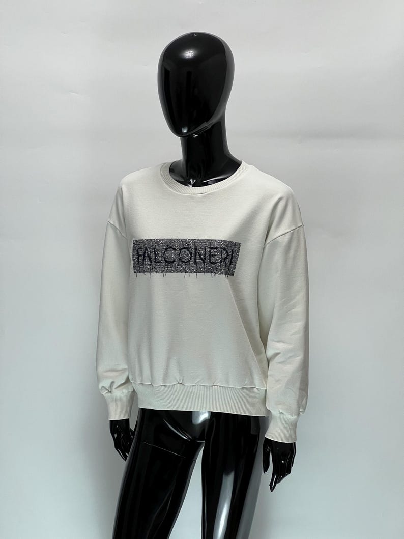 May include: A cream-colored sweatshirt with the word "FALCONERI" in a rectangular design across the chest. The sweatshirt has a crew neck and ribbed cuffs and hem. The garment is displayed on a black mannequin.
