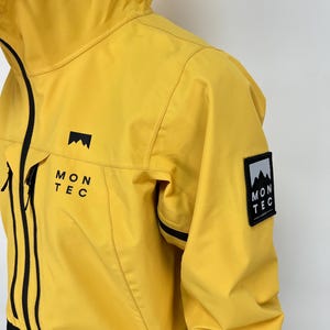 May include: Yellow hooded jacket with black accents and the text "MON TEC". The jacket features a mountain logo on the chest and a patch on the sleeve. The jacket is made of a lightweight material.