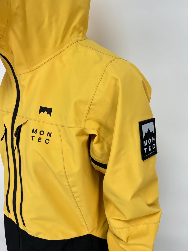 May include: Yellow hooded jacket with black accents and the text "MON TEC". The jacket features a mountain logo on the chest and a patch on the sleeve. The jacket is made of a lightweight material.