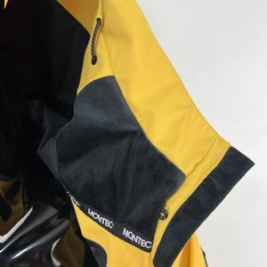 May include: Close-up of a yellow and black jacket. The jacket features a black collar and trim, with the brand name "MONTEC" printed on the collar. The jacket has a drawstring and a glossy black panel.
