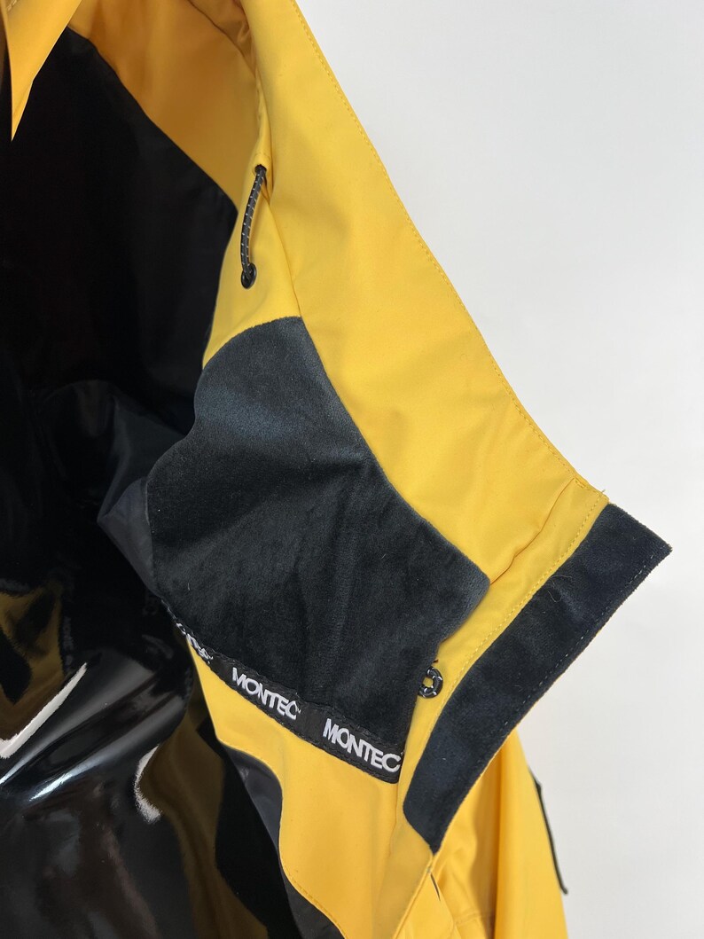 May include: Close-up of a yellow and black jacket. The jacket features a black collar and trim, with the brand name "MONTEC" printed on the collar. The jacket has a drawstring and a glossy black panel.