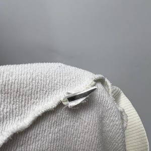 May include: Close-up of a white terry cloth garment, possibly a sweatshirt or towel, with a ribbed collar and a tag. The fabric has a textured, looped surface. The tag is white with a black stripe.