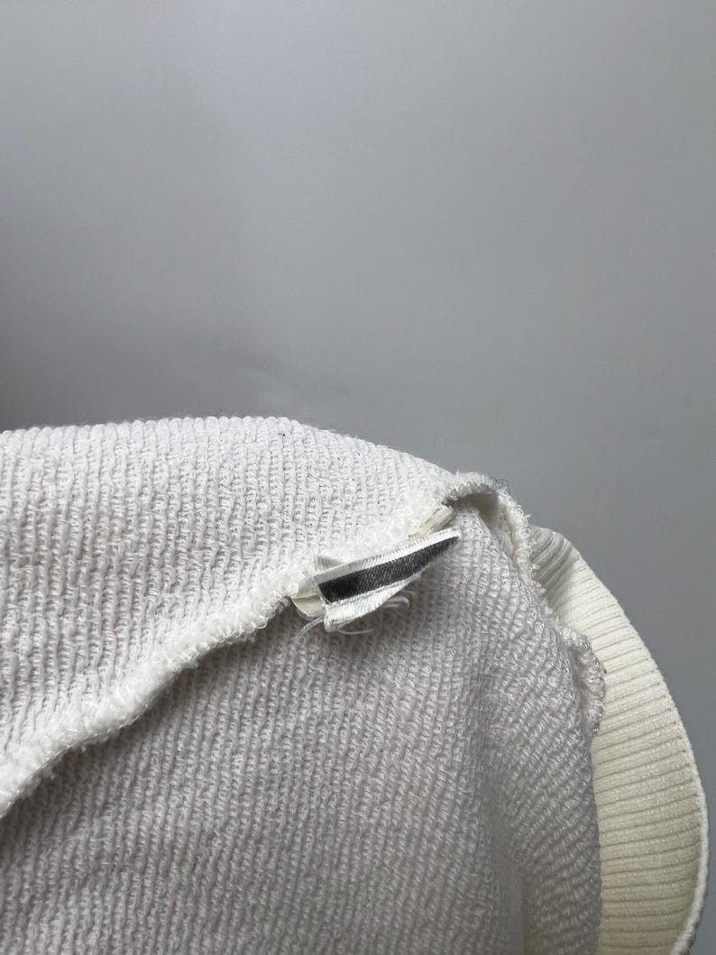May include: Close-up of a white terry cloth garment, possibly a sweatshirt or towel, with a ribbed collar and a tag. The fabric has a textured, looped surface. The tag is white with a black stripe.