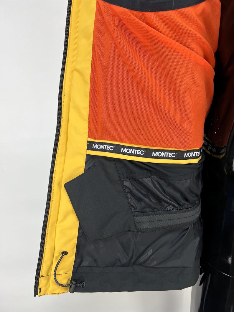 May include: Close-up of a yellow and black winter jacket interior. The jacket features a bright orange lining and a black pocket. The brand name "MONTEC" is printed on a black band. The jacket is designed for cold weather.