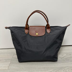 Puede incluir: Un bolso tote negro con asas de cuero marrón y cierre de solapa. El bolso presenta una solapa de cuero marrón con un botón dorado. Las asas son curvas y están unidas a la parte superior del bolso.