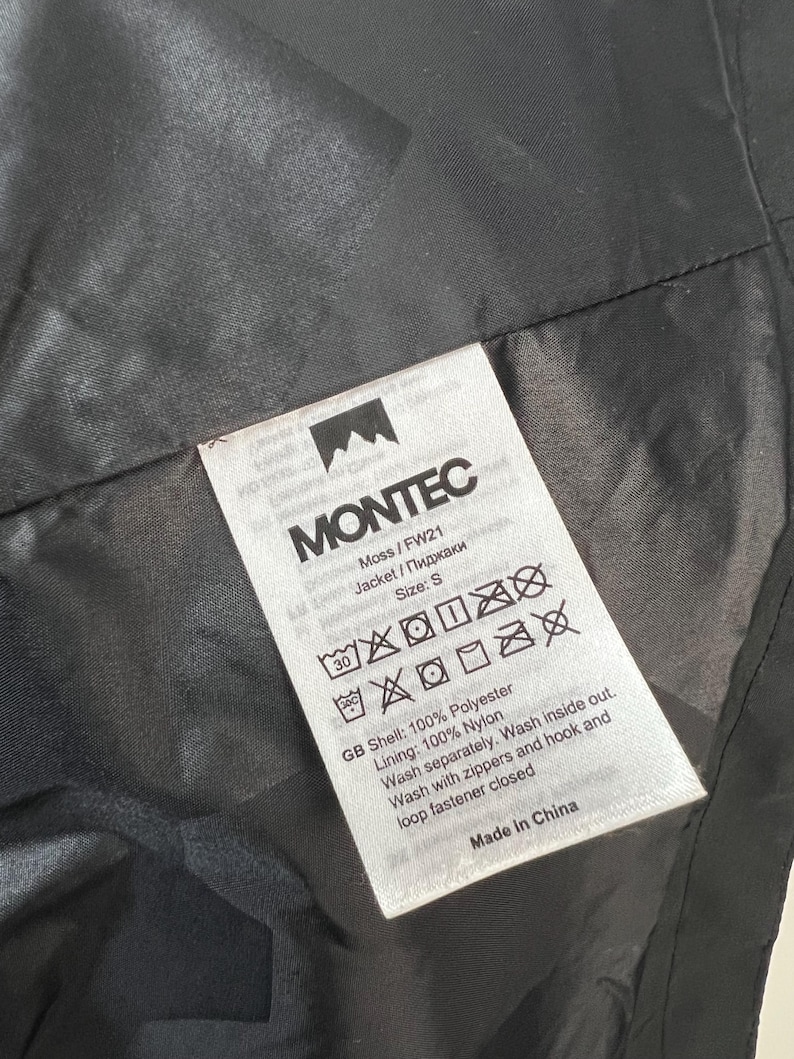 May include: A close-up of a black jacket's interior with a white tag. The tag reads "MONTEC" and includes washing instructions, size "S", and fabric details: 100% polyester shell and 100% nylon lining. The jacket is made in China.
