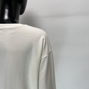 May include: Off-white long-sleeve shirt. The shirt is made of a soft, smooth fabric. The neckline and shoulder seams are visible. The shirt is displayed on a black mannequin.