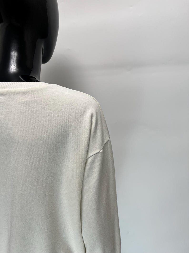 May include: Off-white long-sleeve shirt. The shirt is made of a soft, smooth fabric. The neckline and shoulder seams are visible. The shirt is displayed on a black mannequin.