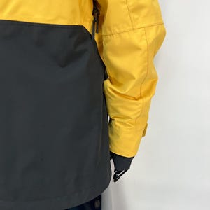 May include: A yellow and black jacket. The upper portion of the jacket is yellow, while the lower portion is black. The jacket has a zipper and a sleeve with a cuff. The jacket is made of a smooth, possibly waterproof material.