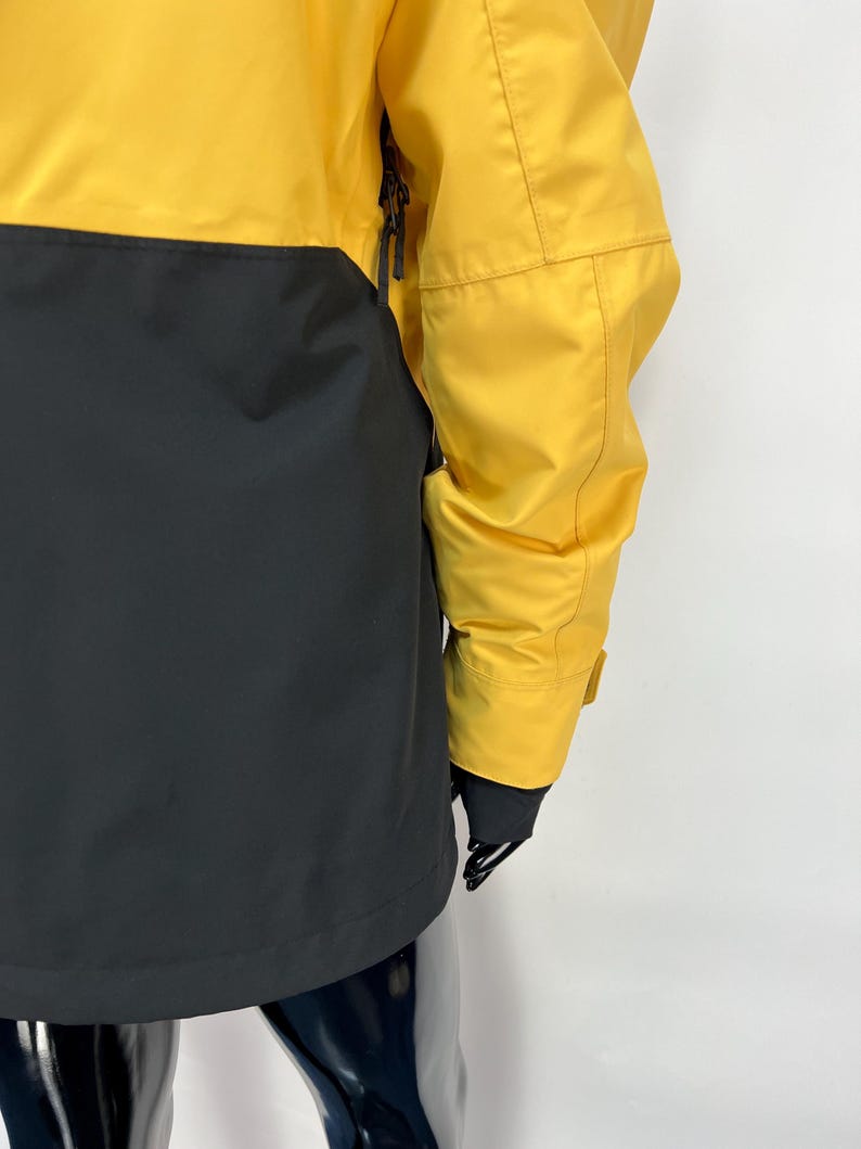 May include: A yellow and black jacket. The upper portion of the jacket is yellow, while the lower portion is black. The jacket has a zipper and a sleeve with a cuff. The jacket is made of a smooth, possibly waterproof material.