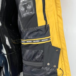 May include: Close-up of a yellow and black winter jacket. The jacket features a black interior with multiple pockets and a yellow exterior. The jacket has the brand name "MONTEC" printed on the interior and a tag with the words "WATERPROOF 20000" and "BREATHABILITY 20000".