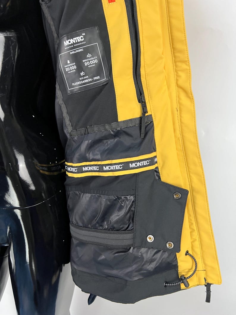 May include: Close-up of a yellow and black winter jacket. The jacket features a black interior with multiple pockets and a yellow exterior. The jacket has the brand name "MONTEC" printed on the interior and a tag with the words "WATERPROOF 20000" and "BREATHABILITY 20000".