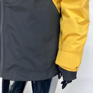 May include: A yellow and black jacket with the word "TEC" embossed on the sleeve cuff. The jacket has a black lower section and yellow upper section. The jacket is made of a smooth, possibly waterproof material.
