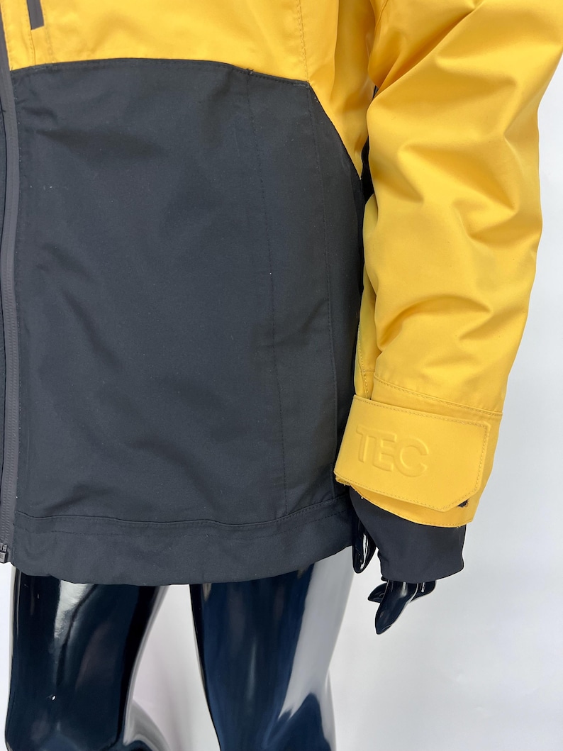 May include: A yellow and black jacket with the word "TEC" embossed on the sleeve cuff. The jacket has a black lower section and yellow upper section. The jacket is made of a smooth, possibly waterproof material.