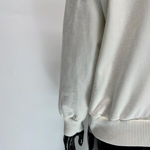 May include: Cream-colored sweatshirt with ribbed cuffs and hem. The long sleeve is gathered at the wrist. The fabric appears soft and slightly textured. The garment is displayed on a black mannequin.