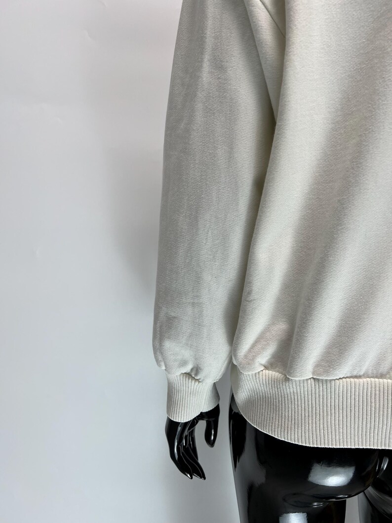 May include: Cream-colored sweatshirt with ribbed cuffs and hem. The long sleeve is gathered at the wrist. The fabric appears soft and slightly textured. The garment is displayed on a black mannequin.