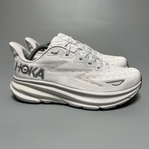 Puede incluir: Un par de zapatillas blancas para correr con la marca "HOKA" en gris en el lateral. Las zapatillas tienen una suela blanca gruesa y curvada y una parte superior de malla transpirable. Los cordones son blancos y el diseño está optimizado para correr o actividades deportivas.