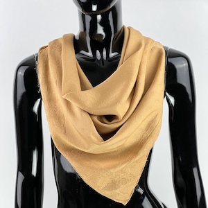 Può includere: Una sciarpa di seta color beige chiaro, drappeggiata attorno al collo di un manichino. La sciarpa ha un motivo sottile e una consistenza morbida e fluida. Di forma quadrata, sembra essere fatta di un materiale leggero.