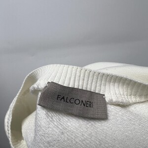 May include: Close-up of a white sweater with a ribbed neckline. A gray label with the brand name "FALCONERI" is sewn on the sweater. The sweater is made of a soft, textured material.